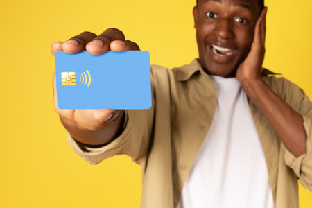 Shocked positive young african american guy in casual presses hand to cheek, shows credit cardの写真素材