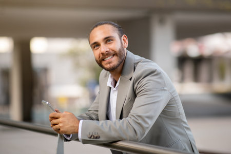 Pensive Young Businessman With Smartphone In Hands Standing Outdoors And Looking Awayの写真素材