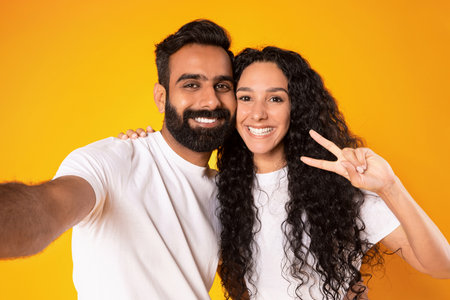 Arab couple posing for selfie gesturing victory sign, yellow background
