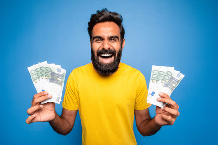 Lucky winner. Euphoric middle aged indian man holding euro money cash in hands, posing on blue...