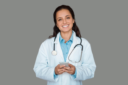 Doctor woman holding smartphone using medical application, standing on grey studio background, smiling at cameraの写真素材