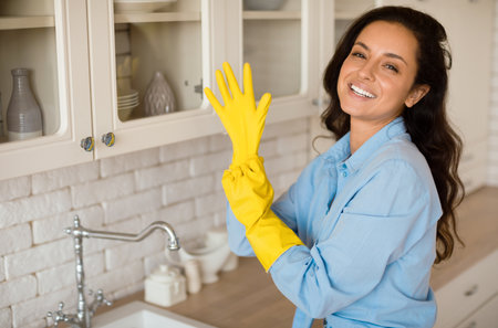 Portrait of joyful woman putting on rubber gloves, ready to tidy house or washing dishes standing in...