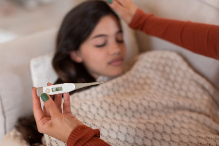 Sick girl with fever lying while mom holding thermometer indoorの写真素材