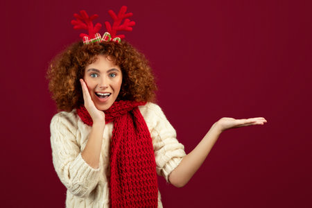 Happy surprised teen caucasian curly girl with deer antlers, scarf with open mouth hold empty space on handの写真素材