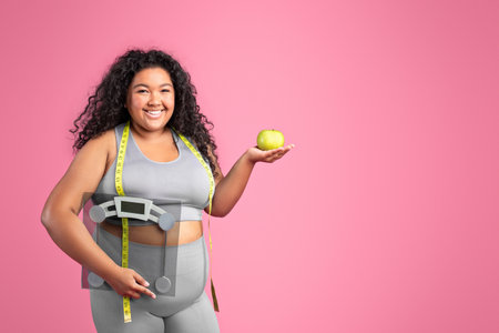 Glad black body positive woman in sportswear holding apple and scales, enjoying diet result, pink...