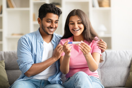 Excited indian couple looking at positive pregnancy test, smiling and enjoying great news, sitting on couch at homeの写真素材