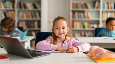 Smiling european schoolgirl at desk with laptop, writing in copybookの写真素材