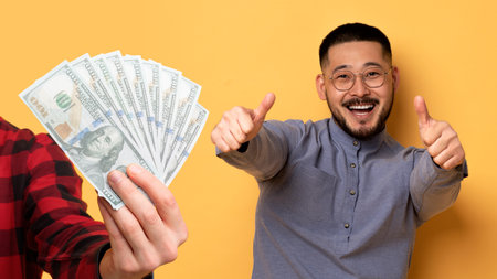 Cheerful millennial chinese guy pointing thumbs up, near hand with many money