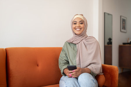 Woman In Hijab Holding Smartphone Sitting On Sofa At Home