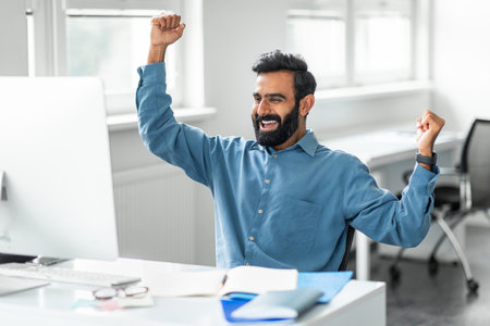 Indian businessman looking at computer monitor and shaking fists celebrating big luck in officeの写真素材