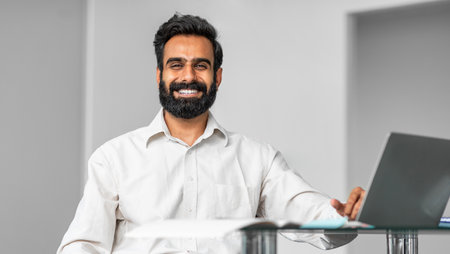 Portrait of happy indian man sitting at desk in office, working on laptop and smiling at camera, panorama, free spaceの写真素材