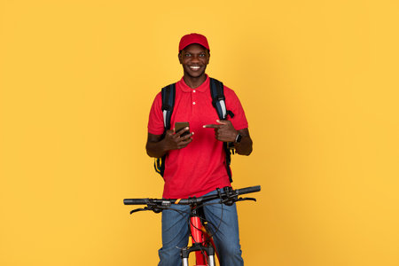 Cheerful millennial black man courier in uniform with backpack on bike, point finger at phone