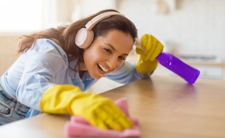 Joyful woman cleaning table with spray, headphones onの写真素材