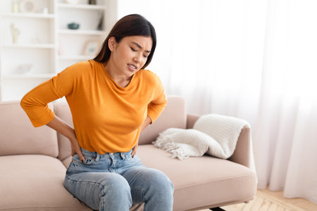 Unhappy asian woman sitting on couch, touching back, have painの写真素材