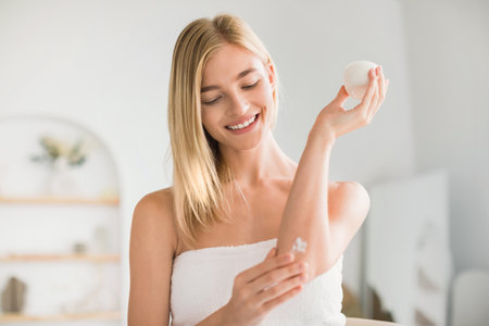 Happy Blonde Lady Applying Moisturizer On Elbow At Home Bathroomの写真素材