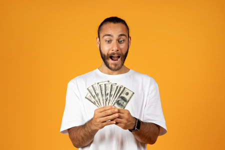 Happy shocked millennial caucasian man in white t-shirt with open mouth look at many money dollars in handsの写真素材