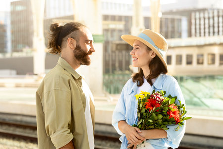 Positive young caucasian husband meets surprised lady, gives bouquet of flowers on train stationの写真素材