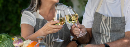 Cheerful senior caucasian man and lady in aprons cheers glasses of wine at table in home gardenの写真素材