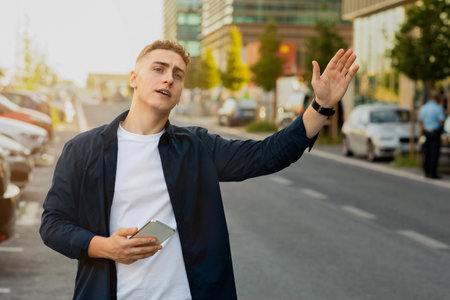 Serious handsome millennial caucasian man in casual, use smartphone in city street, stop taxi with handの写真素材