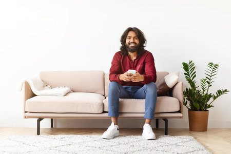 Positive indian man sitting on couch and using cell phoneの写真素材
