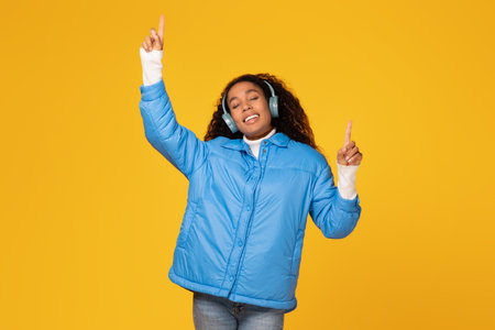 Happy black lady in jacket enjoys music on yellow backgroundの写真素材