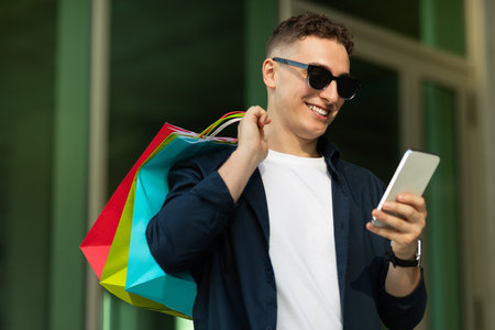 Positive young caucasian man in casual, sunglass, with many bags, chatting by mobile phone, near city mallの写真素材