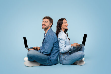 Couple sitting back-to-back with laptops, blue backgroundの写真素材