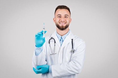 Cheerful young doctor man in white coat and gloves with syringe, grey backdropの写真素材