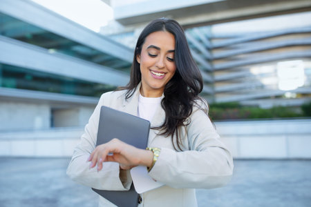 Latin entrepreneur woman smiling checking watch standing with laptop outdoorの写真素材