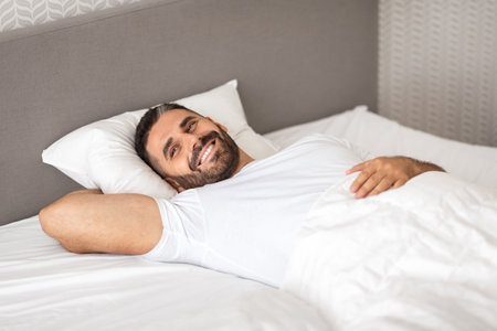 Relaxed European man happily lounging in comfortable bed in bedroomの写真素材