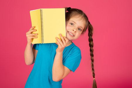 Cheerful young girl with braids holding yellow notepad and smiling at cameraの写真素材