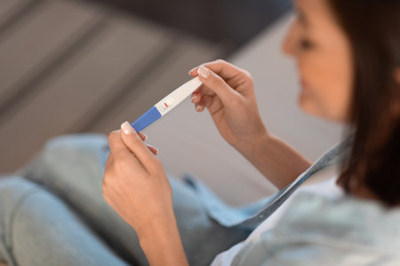 Smiling pregnant lady holds positive pregnancy test sitting indoor, cropped