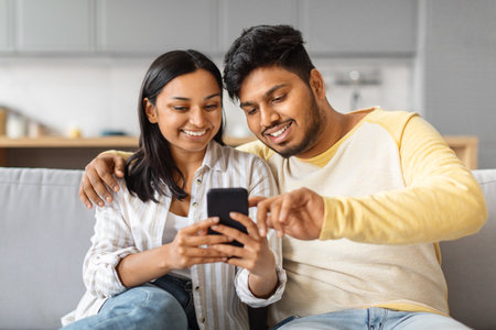 Smiling Young Indian Couple Using Smartphone And Embracing Together At Homeの写真素材