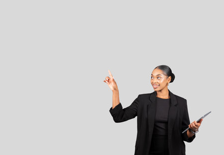 Smiling African American businesswoman in professional attire holding a tablet while pointing upwardsの写真素材