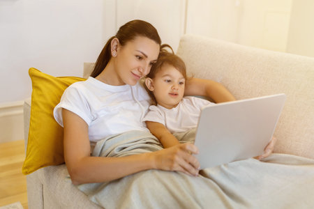 Mother and little daughter with digital tablet relaxing under blanket on sofaの写真素材