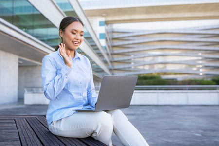 Japanese woman sitting outdoors with laptop video calling waving handの写真素材
