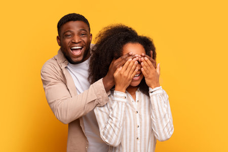 Black man covering womans eyes for surprise on yellow backgroundの写真素材