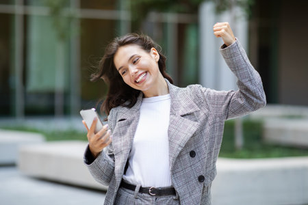 Joyful businesswoman using phone and shaking fist in celebration outdoor