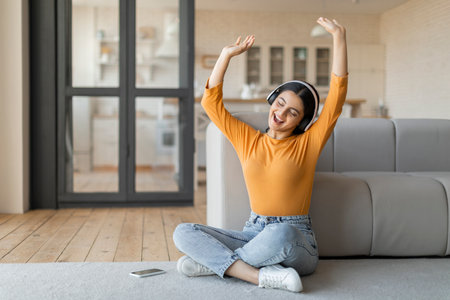 Joyful young indian woman wearing headphones listening music on smartphone at homeの写真素材