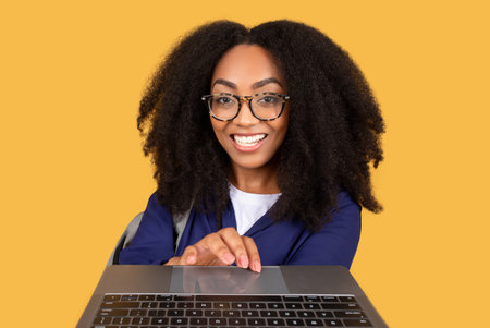 Cheerful black lady student typing at keyboard laptop and smiling on yellow backgroundの写真素材