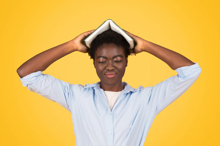 Tired teen black woman student hold book on head, joyful expression with closed eyes