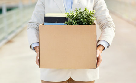 A person holding a cardboard box filled with office supplies and a green plantの写真素材