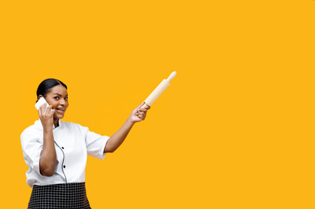 Efficient black female chef managing tasks over phone and holding rolling pinの写真素材