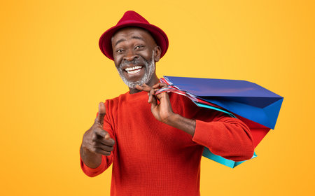 Charismatic senior Black man with a white beard, giving a thumbs up and carrying colorful shopping bagsの写真素材