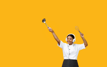 Black female chef wearing wireless headphones lifting cooking utensilsの写真素材