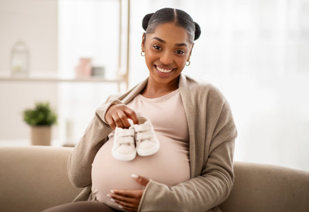 Pregnant black woman holding small baby shoes relaxing at homeの写真素材