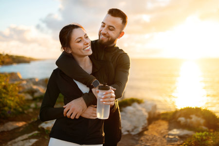 Loving couple embracing at sunset, woman with water bottle, man hugging her from behind, sunlit sea in backgroundの写真素材