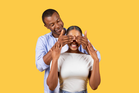 Black man covering womans eyes with hands, both smiling on yellowの写真素材