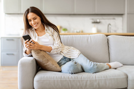 Contented female enjoying browsing her smartphone, sitting on couch in living roomの写真素材