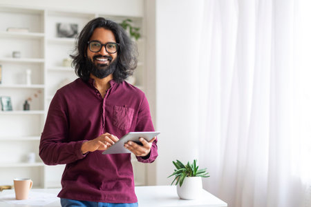 Handsome Indian Man With Digital Tablet Standing Near Desk At Homeの写真素材